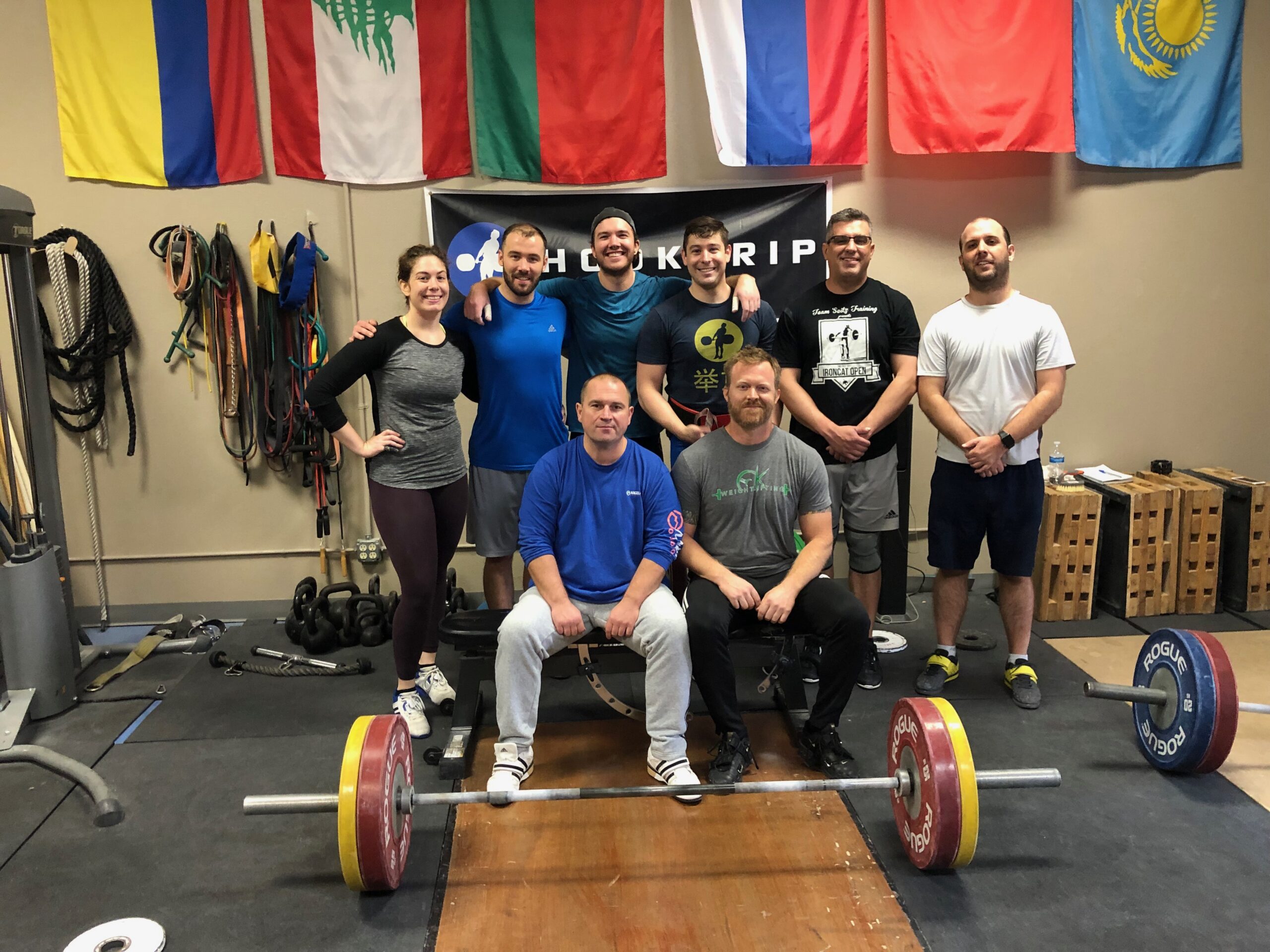 Group of olympic weightlifters posing for a group photo in front of a barbell with flags behind them
